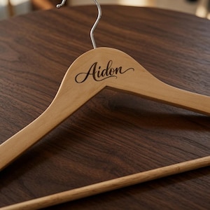May include: A light-colored wooden clothes hanger featuring a silver metal hook at the top. The wood surface is engraved with the name Aidon in a dark, elegant, cursive script font, positioned centrally on the upper part of the hanger frame.
