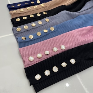 May include: A stack of long-sleeved shirts in various colors, each with a row of gold buttons down the sleeve. The buttons are either round or oval shaped.