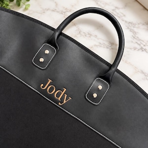 May include: A black garment bag featuring a smooth upper panel and a textured lower panel, separated by white stitching. The name Jody is embroidered in gold thread. It includes two black handles attached with silver-toned metal rivets.