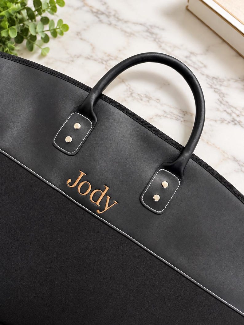 May include: A black garment bag featuring a smooth upper panel and a textured lower panel, separated by white stitching. The name Jody is embroidered in gold thread. It includes two black handles attached with silver-toned metal rivets.