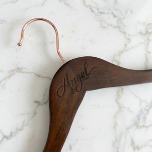 May include: A dark brown wooden clothes hanger featuring a rose gold metal hook. The word Angel is engraved in a cursive script font on the upper left side of the wooden frame of the hanger, showing a smooth, polished finish.