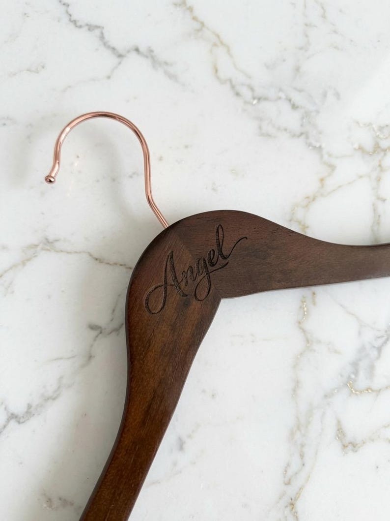 May include: A dark brown wooden clothes hanger featuring a rose gold metal hook. The word Angel is engraved in a cursive script font on the upper left side of the wooden frame of the hanger, showing a smooth, polished finish.