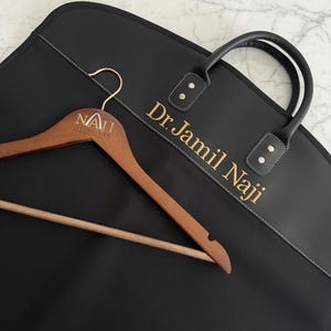 May include: A black garment bag with the text Dr. Jamil Naji printed in gold, featuring black handles and a wooden clothes hanger with the engraved text NAJI INJURY LAW, positioned diagonally across the front of the bag.