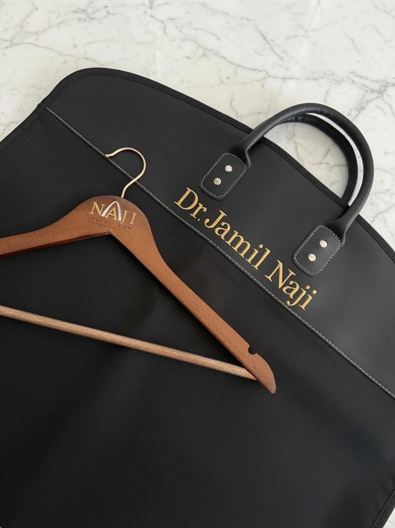 May include: A black garment bag with the text Dr. Jamil Naji printed in gold, featuring black handles and a wooden clothes hanger with the engraved text NAJI INJURY LAW, positioned diagonally across the front of the bag.