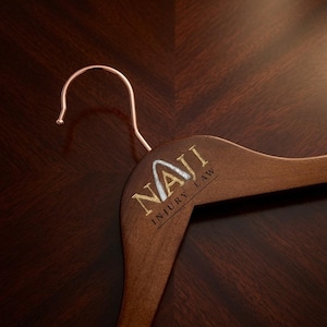May include: A dark brown wooden clothes hanger with a rose gold metal hook. The hanger features the gold and silver text NAJI INJURY LAW printed on the front, with the letter A stylized to resemble a mountain peak or upward arrow.