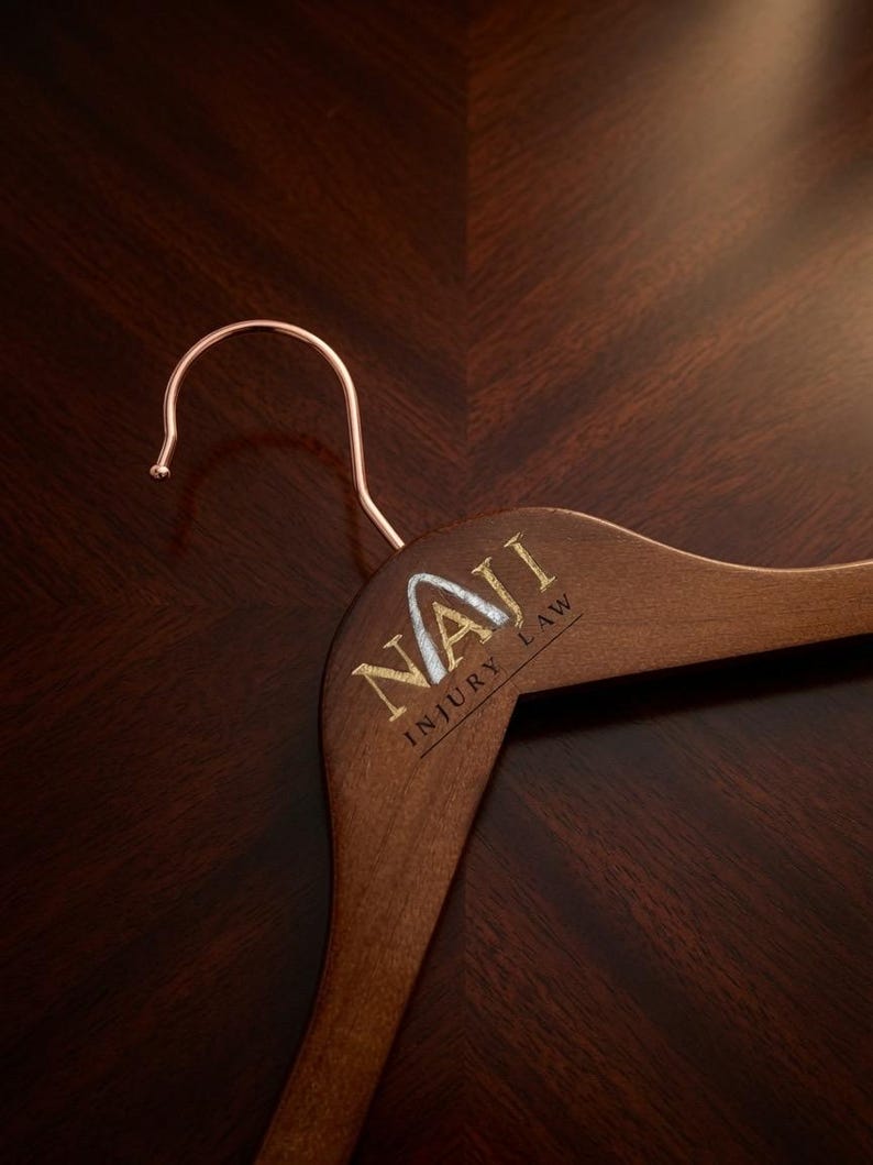 May include: A dark brown wooden clothes hanger with a rose gold metal hook. The hanger features the gold and silver text NAJI INJURY LAW printed on the front, with the letter A stylized to resemble a mountain peak or upward arrow.