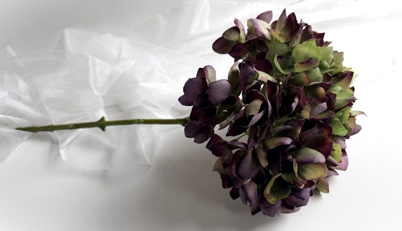 May include: A single stem of artificial hydrangea flowers. The flower head is a mix of deep purple and green hues, with a long green stem. The background is white with a translucent, crinkled material.