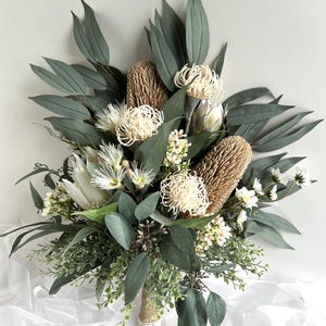 Pode incluir: Um exuberante buquê com uma variedade de flores e folhagens artificiais. O arranjo inclui grandes flores texturizadas marrons e creme, cercadas por folhas verdes escuras e flores brancas menores. As hastes são embrulhadas em juta e renda.