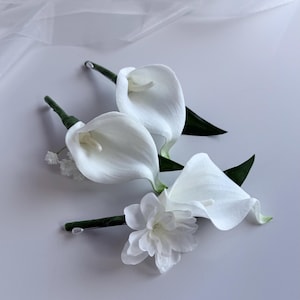 May include: Three white calla lilies and a white flower with green stems and leaves are arranged on a white surface. The calla lilies have a smooth, elegant shape, and the other flower has multiple petals.
