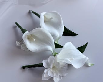 May include: Three white calla lilies and a white flower with green stems and leaves are arranged on a white surface. The calla lilies have a smooth, elegant shape, and the other flower has multiple petals.