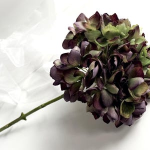 May include: A single stem of artificial hydrangea with a large, full bloom. The flower petals are a mix of deep purple, burgundy, and green hues. The stem is a light green color, and the flower is set against a white background.