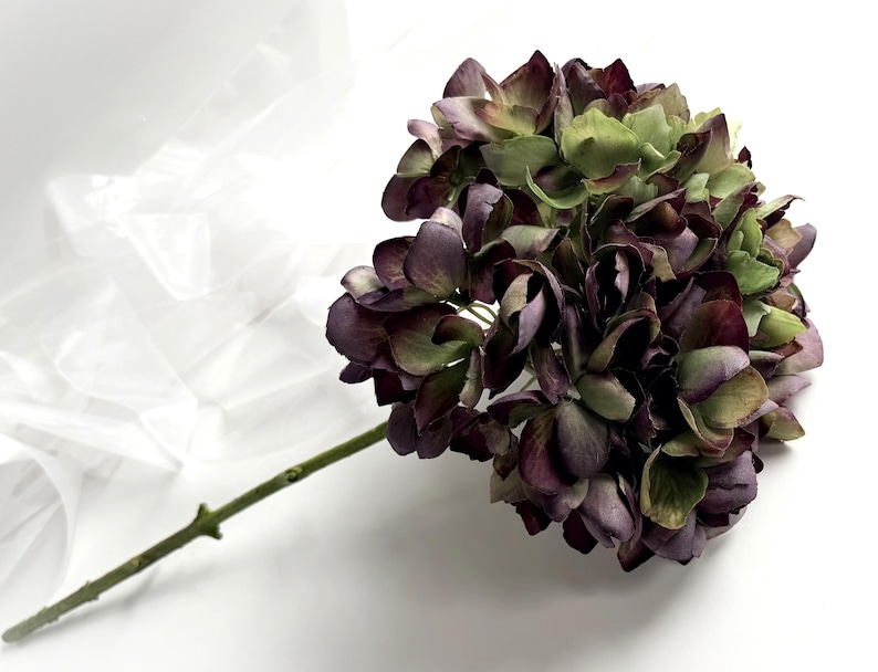 May include: A single stem of artificial hydrangea with a large, full bloom. The flower petals are a mix of deep purple, burgundy, and green hues. The stem is a light green color, and the flower is set against a white background.