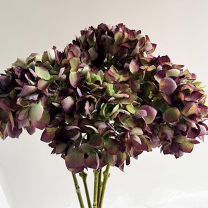 May include: A bouquet of faux hydrangeas in shades of deep purple, green, and burgundy. The artificial flowers have multiple layers of petals and are clustered together, with several green stems visible at the base. The arrangement is set against a white background.