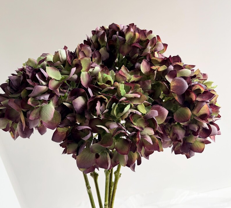 May include: A bouquet of faux hydrangeas in shades of deep purple, green, and burgundy. The artificial flowers have multiple layers of petals and are clustered together, with several green stems visible at the base. The arrangement is set against a white background.