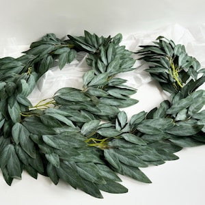 May include: Two dark green leaf garlands arranged in a circular shape. The leaves are a muted green colour with a slightly textured appearance. The garlands are likely used for home decor or crafting.