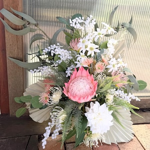 May include: A vibrant floral arrangement featuring a large pink protea flower as the focal point, surrounded by white, pink, and green foliage. The bouquet includes white dahlias, delicate white flowers, and textured green leaves, creating a lush, natural display.