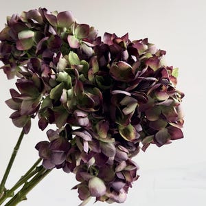 May include: A close-up of a bouquet of faux hydrangea flowers. The flowers are a mix of deep purple, green, and cream colors. The stems are green and the background is white.