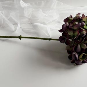 May include: A single artificial flower with a long green stem and a cluster of dark purple and green petals. The flower is set against a white background with a sheer, white fabric in the upper left corner. The flower's stem is approximately 12 inches long.