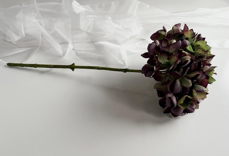 May include: A single artificial flower with a long green stem and a cluster of dark purple and green petals. The flower is set against a white background with a sheer, white fabric in the upper left corner. The flower's stem is approximately 12 inches long.