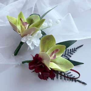 May include: Two floral boutonnieres featuring light green cymbidium orchids. One boutonniere includes white blossoms and green leaves, while the other features deep burgundy petals, dark green fern-like foliage, and a thin burgundy loop.