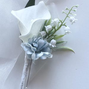 May include: A floral boutonniere featuring a single white calla lily, a small dusty blue carnation, and a sprig of white lily of the valley. The stem is wrapped in white satin ribbon and includes a silver pin with a small white pearl-like bead.