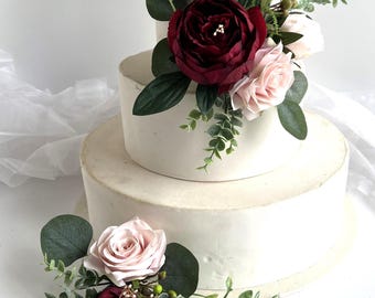 May include: A three-tiered white cake decorated with burgundy and blush-coloured flowers and greenery. The cake is round and has a smooth, frosted finish. The floral arrangement includes roses and other decorative foliage.