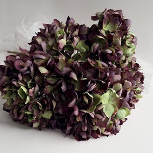 May include: A bouquet of artificial hydrangeas in shades of deep purple and green. The flowers have a realistic appearance with layered petals. A sheer white fabric is visible in the background, adding a soft contrast to the bouquet.