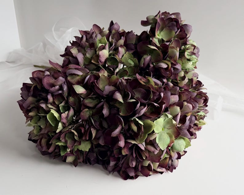 May include: A bouquet of artificial hydrangeas in shades of deep purple and green. The flowers have a realistic appearance with layered petals. A sheer white fabric is visible in the background, adding a soft contrast to the bouquet.