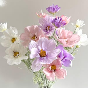 Könnte beinhalten: Ein Strauß künstlicher Kosmosblumen in Weiß, Rosa und Lila. Die Blumen haben gelbe Zentren und grüne Stiele. Die Anordnung befindet sich in einer durchsichtigen Glasvase.