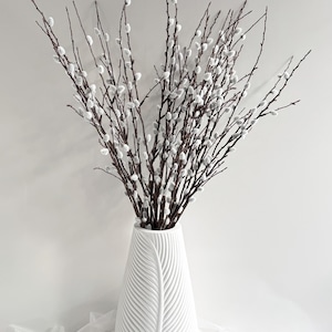 May include: A white ceramic vase with a leaf design holds a bundle of pussy willow branches. The branches have dark brown stems and are dotted with small, white, fluffy catkins. The vase has a textured surface and a wide base.