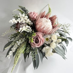 May include: A lush bouquet featuring a large pink protea flower as the focal point, surrounded by dusty rose roses, white and green foliage, and accents of white flowers. The arrangement is set against a white backdrop.