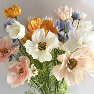 Könnte beinhalten: Ein lebendiger Strauß künstlicher Blumen in verschiedenen Farben, darunter Weiß, Pfirsich, Orange, Gelb und Blau. Das Arrangement zeigt eine Mischung aus verschiedenen Blumensorten mit grünem Laub, wodurch eine farbenfrohe und fröhliche Darstellung entsteht.
