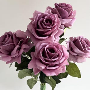 May include: A bunch of five mauve roses with green foliage. The rose heads are fully open, displaying their layered texture. The stems are visible, bound with a green ribbon.