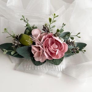 Puede incluir: Un peine para el cabello floral con una gran peonía rosa, flores rosas más pequeñas y hojas verdes. El peine incluye ramitas de vegetación y una vaina verde. Las flores están unidas a un peine de plástico transparente.
