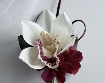 May include: A floral boutonniere featuring a large white orchid with a deep burgundy center, accented by smaller dark red petals, green leaves, and a thin dark stem. The stem is wrapped in green tape and finished with a small white pearl pin.