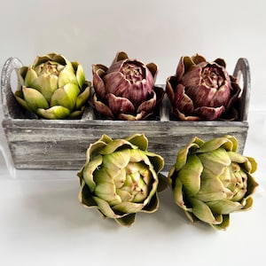 May include: A grey wooden crate contains three artichokes, one green and two burgundy. Two more green artichokes are in front of the crate. The artichokes have layered petals and are on a white surface.