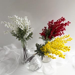 May include: Three glass vases display colourful floral arrangements. One vase holds white flowers, another red, and a third yellow. The vases are clear and sit on a white surface, with a sheer white fabric in the background.