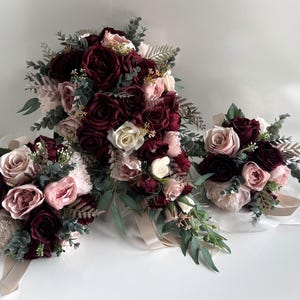 May include: Three bouquets of artificial flowers in burgundy, blush pink, and cream. The bouquets feature roses, carnations, and greenery, with satin ribbons. The largest bouquet is a cascading style, the others are round.