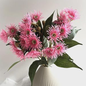 以下が含まれることがあります： 白い花瓶に入った、とげのある花びらと緑の葉を持つピンクの花のブーケ。 花には中央に茶色の芯があり、ふっくらと豊かに配置されています。 花瓶はリブ編みのテクスチャです。
