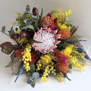 May include: A colourful bouquet with a large, pale pink protea flower at its centre, complemented by yellow, orange, and red blooms, green leaves, and dark brown seed pods. The arrangement is displayed against a white backdrop.
