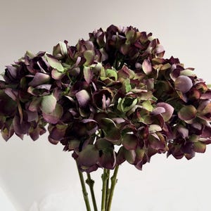 May include: A bouquet of artificial hydrangeas in shades of deep purple, green, and brown. The flowers have multiple layers of petals and are clustered together. The stems are a light brown color, and the background is a plain white.