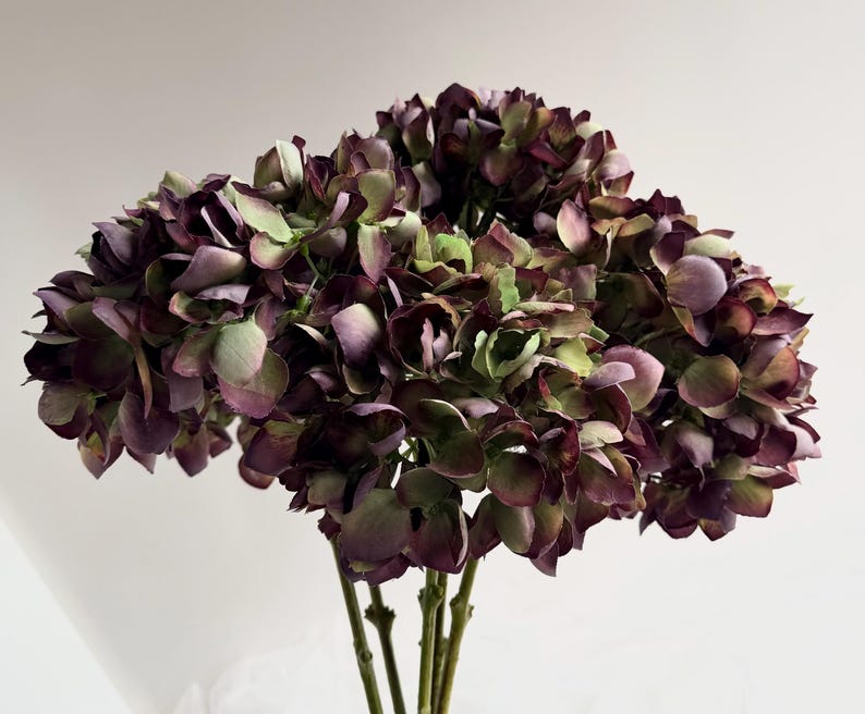 May include: A bouquet of artificial hydrangeas in shades of deep purple, green, and brown. The flowers have multiple layers of petals and are clustered together. The stems are a light brown color, and the background is a plain white.