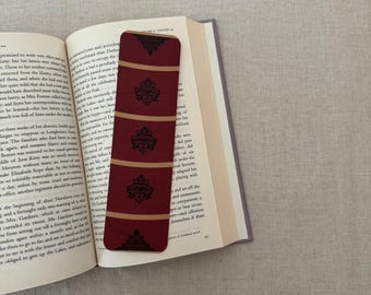 May include: A burgundy bookmark with a repeating gold and black damask pattern is placed in an open book. The bookmark has a rectangular shape with rounded corners. The book's pages are off-white.