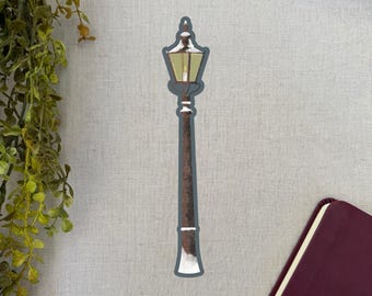 May include: A sticker featuring a vintage-style street lamp. The lamp has a brown pole with a dark teal outline and a yellow light. The top of the lamp and the base have white accents, suggesting snow. The sticker is on a light gray background.