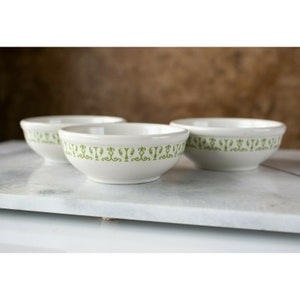May include: Three white ceramic bowls with a green decorative pattern around the rim. The bowls are arranged in a row on a white marble surface.