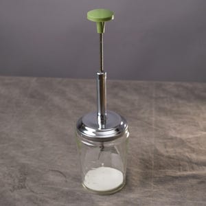May include: A vintage glass sugar dispenser with a chrome lid and a green plastic handle. The dispenser is filled with white sugar. The design is simple and functional, typical of mid-century kitchenware.