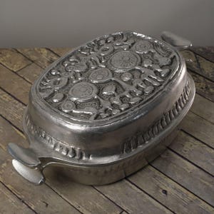 May include: A silver-colored, oval-shaped decorative dish with a detailed, raised floral design on the lid. The dish has two handles and a textured border. The dish is sitting on a wooden surface.