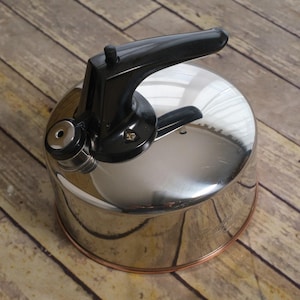 May include: A polished stainless steel tea kettle with a cylindrical body and a copper-toned base. The kettle features a black plastic handle with a thumb-operated lever mechanism and a small circular steam vent located near the spout assembly.