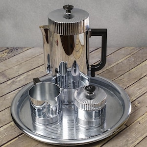 May include: A chrome coffee set with a ribbed design, including a coffee pot, creamer, sugar bowl, and a tray. The coffee pot has a black handle and a black knob on the lid. The set is displayed on a wooden surface.