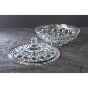 May include: A clear glass serving bowl with a matching lid. The bowl has a ribbed design around the rim and a diamond pattern on the base. The lid features a diamond pattern and a small knob handle. The bowl and lid are on a gray surface.